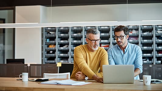 Two businessmen behind a desk. The desk has a laptop, two cups of coffee and papers on it.
